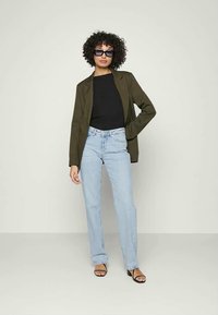 Olivově zelený blazer přes černý top, spárovaný s světle modrými rovnými džíny a černými sandály na podpatku. Modelka s kudrnatými vlasy a slunečními brýlemi.