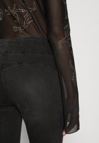 Top noir transparent avec des manches longues et des embellissements argentés. Jean noir taille haute avec une texture lisse et des détails brodés.