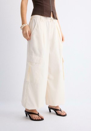 Osoba nosiaca voľné béžové cargo nohavice s širokými nohavicami, čierne sandále na podpätku, hnedý skrátený top a náramok s nitmi na ľavom zápästí.