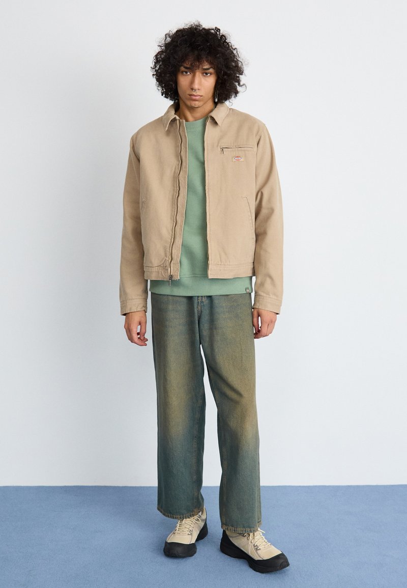 Beige bomullsjacka med dragkedja och vänster bröstficka, lager på lager över en grön sweatshirt. Blågröna jeans och beige skor fulländar looken.