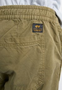 Shorts cargo verts olive avec une taille élastique et une poche latérale. Présente une étiquette noire avec du texte jaune de Alpha Industries.