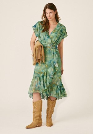 Mujer con vestido cruzado verde con estampado floral y volantes, botas de vaquero marrones y sosteniendo un bolso marrón con flecos sobre un fondo liso.