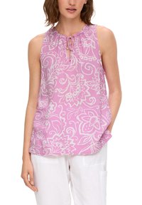 Blusa senza maniche rosa con un motivo floreale bianco, scollo arricciato con lacci e una vestibilità ampia e fluida. Indossata con pantaloni bianchi.