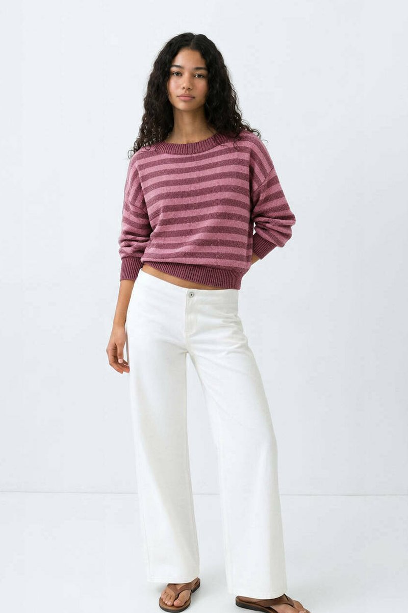 Femme aux cheveux bouclés portant un pull rayé mauve, un pantalon blanc à jambes larges et des sandales marron, debout devant un fond blanc uni.