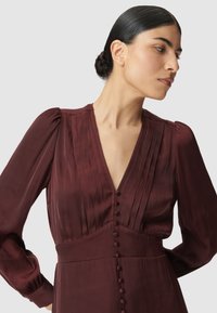 Robe bordeaux avec des détails plissés, décolleté en V profond, manches bouffantes longues et accents de boutons sur le devant. Tissu lisse et texturé.