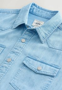 Chemise en denim bleu clair avec boutons pression en métal, poche poitrine à rabat et col pointu, étalée à plat sur une surface blanche.