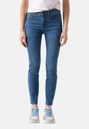 Kobieta ubrana w niebieskie rurki z średnim stanem i jasnoniebieskie sportowe buty z różowymi akcentami, stojąca na jednolitym tle.