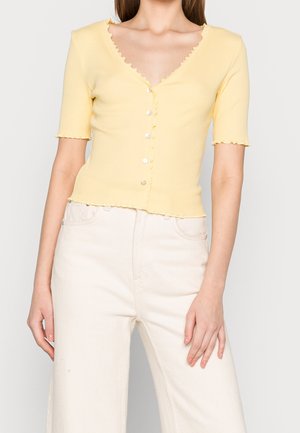 Kobieta ubrana w bluzkę na guziki o krótkich rękawach w bladym żółtym kolorze z ozdobnymi falbankami oraz w szerokie, wysokiej talii spodnie w kolorze kości słoniowej.