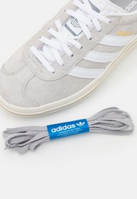 Gray suede sneaker with white stripes, flat white laces, and an off-white sole. Accompanied by gray laces with a blue Adidas label.