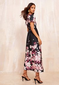 Ziedu maxi kleita melnā satinā ar rozā un baltām puķēm, īsām pūkainām piedurknēm, pieguļošu vidukli un plūstošu siluetu. Savienota ar melniem apavu papēžiem ar siksnām.