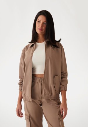Beige bomberi takki, jossa on vetoketju edessä, joustavat hihansuut ja pieni logotyyppi. Päällä valkoinen ribbattu crop-top. Sujuva pinta.