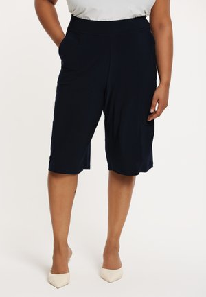 Marineblaue, knielange Shorts mit glatter Textur, breitem Bund und seitlichen Taschen, kombiniert mit beigen spitz zulaufenden Schuhen.