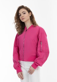 Giacca bomber rosa con chiusura a zip, polsini a coste e tasca laterale. Texture liscia e design cropped, indossata sopra pantaloni bianchi.