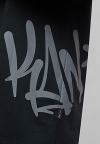 Black t-shirt featuring large gray graphic text design. Soft cotton material with a relaxed fit and visible brand tag at the side.