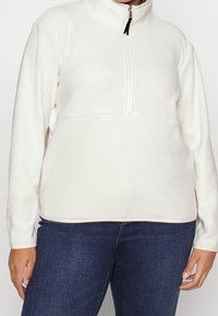 Valkoinen fleece-pusero, jossa on puolivetoketju-kaulus, sileä pinta ja pitkät hihat. Huomattava ompeluseikka rinnan sauman kohdalla.