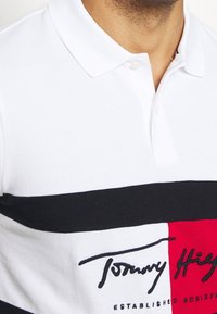 Polo blanc en tissu texturé avec des rayures horizontales noires et rouges, et un logo avec du texte signature sur le devant.