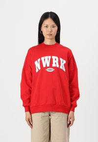 Felpa rossa con "NWRK" in bold lettere bianche sulla parte anteriore, colletto e polsini a costine. Tessuto morbido con vestibilità comoda.