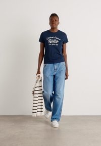 Marineblå kortermet T-skjorte med hvitt logo, sammen med lyseblå vide jeans. Modellen holder en stripete genser i hvitt og marineblått. Hvite sneakers.