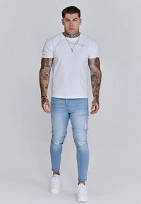 Fehér rövid ujjú póló, kék, kopott hatású szűk farmernadrág és fehér sportcipő. Tetoválások láthatók a karokon, világos háttér előtt állva.