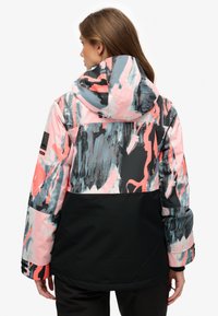 Chaqueta impermeable colorida con una mezcla de patrones abstractos en rosa, gris y negro, con capucha y sección inferior negra contrastante.