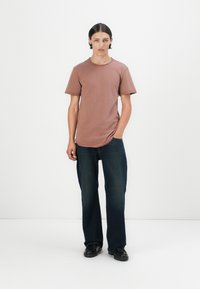 Rostfarbenes Baumwoll-T-Shirt mit Rundhalsausschnitt und kurzen Ärmeln, kombiniert mit dunkelblauen, geraden Jeans und schwarzen Schuhen.