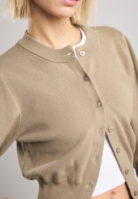Beige ribbed cardigan with a round neckline, featuring button closure and gathered hem detail, layered over a white top.