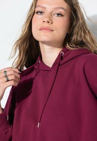 Joven mujer con ojos claros y pelo suelto ajustando el cordón de su sudadera color burdeos, luciendo anillos de plata en sus dedos.
