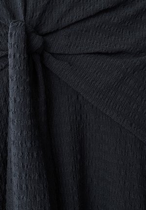 Tejido negro con textura y superficie arrugada, que presenta un detalle anudado. El diseño incluye pliegues drapeados y un acabado suave y desigual.