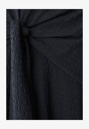 Tejido negro con textura y superficie arrugada, que presenta un detalle anudado. El diseño incluye pliegues drapeados y un acabado suave y desigual.