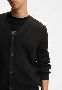 Svart cardigan med en strukturerad, ribbad design, som har en V-halsringning, fem knappar och uppvikt mudd. Diskret logotyp vid fållen.