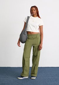 Ung kvinne med lange fletter har på seg en hvit cropped t-skjorte, grønne vide bukser, marineblå sneakers, og bærer en grå skulderveske mot en hvit vegg.