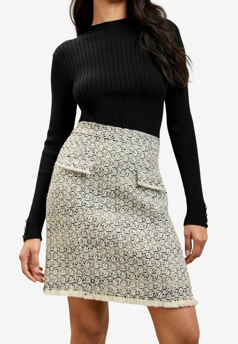 Femme portant un haut noir à manches longues côtelé et une jupe taille haute en tweed blanc avec poches à rabat sur le devant et ourlet effiloché.