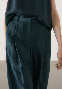 Dark teal linen trousers with a relaxed fit, featuring a flat front, pleats, and a matching belt, shown with a textured top.