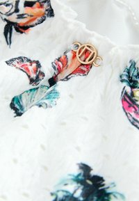 Țesătură albă cu motive florale colorate, suprafață texturată și un detaliu hardware circular auriu. Prezintă un guler strâns cu perforații.