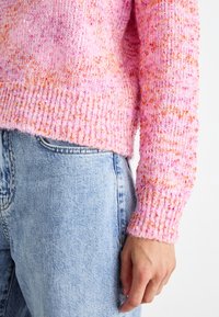 Pletený svetr se světle růžovým přechodem a oranžovými tečkami. Žebrované manžety kontrastují s hladkou, měkkou texturou. Nošený s světle modrými džínami.