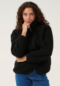 Svart fleecejacka med hög krage, dragkedjestängning och mjukt texturerat tyg. Bärs över blå denimjeans med avspänd passform och rundad nederkant.