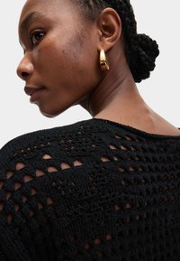 Pull en tricot noir avec un motif en dentelle ajouré et un encolure plongeante ; boucle d'oreille en or visible en gros plan sur le profil du modèle.