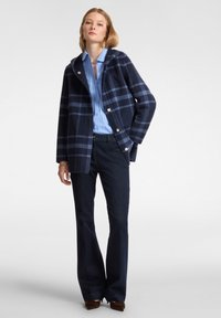 Abrigo de mezcla de lana azul marino con un gran patrón de cuadros azules y negros, capucha y cierre con botones plateados, combinado con una camisa azul clara y jeans oscuros.