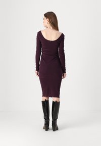 Long-sleeve, form-fitting dress in deep purple fabric with a low scoop back, paired with glossy black knee-high boots.
