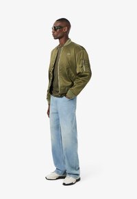 Chaqueta bomber verde oliva con cierre frontal, puños de canalé y detalles texturizados en las mangas, combinada con jeans azul claro y zapatillas blancas.