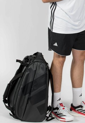 Person trägt schwarze Shorts, weißes Sportshirt, weiße Adidas-Socken und Turnschuhe und steht neben einem großen schwarzen Sport-Rucksack vor einem einfarbigen Hintergrund.
