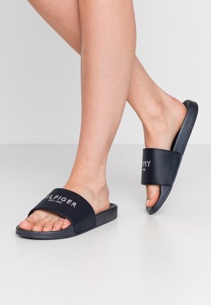 Mørkeblå glidesandaler med en bred rem med hvid 'TOMMY HILFIGER' logo. Tekstureret gummisål og åben tå-design.