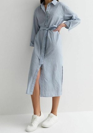 Nainen, joka on pukeutunut sinivalkoraidalliseen midimekkoon, jossa on napit edessä ja vyötäröllä solmittu nauha, sekä valkoiset tennarit, seisoo yksinkertaista vaaleaa seinää vasten.