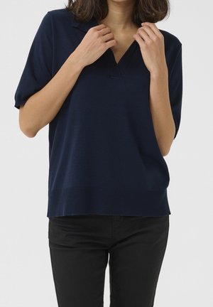 Femme ajustant le col d’un pull bleu marine à manches courtes et col en V, portant un pantalon noir sur un fond uni.