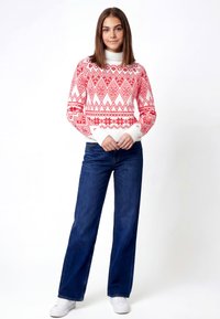 Suéter blanco con patrones geométricos rojos, cuello alto y puños acanalados, combinado con pantalones vaqueros azules de pierna ancha y zapatillas blancas.