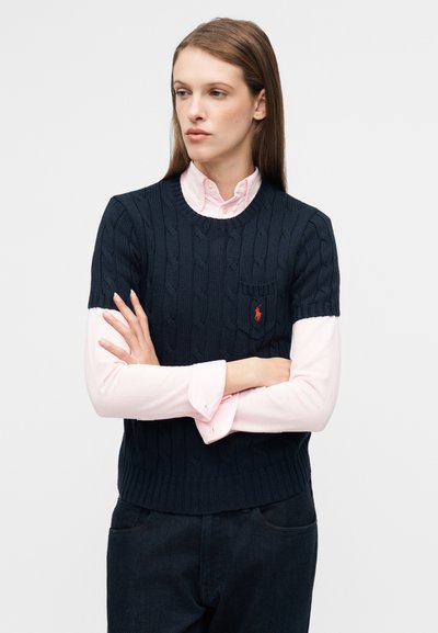 Tummansininen palmikkoneulepaita pyöreällä kaula-aukolla, lyhyillä hihoilla ja rintataskulla; kerroksittain vaaleanpunaisen nappikauluspaidan päällä.