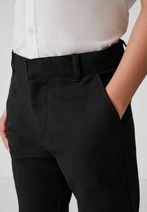 Person wearing black pants with hand in pocket and a tucked-in white button-up shirt on a plain background.