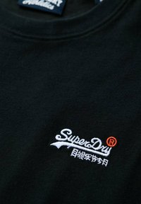 Camiseta de algodón negra con cuello redondo, que presenta un logotipo bordado de "SuperDry" en blanco y naranja, junto con un detalle de texto adicional.