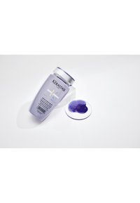 Lavendel-Shampoo-Flasche mit der Aufschrift Kérastase Blond Absolu, schräg auf einer weißen Oberfläche liegend mit einer kleinen Pfütze lila Flüssigkeit daneben.
