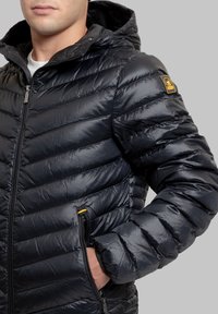 Giacca imbottita nera con finitura lucida, cuciture orizzontali, cappuccio e tasche con cerniera. Presenta un piccolo logo sulla manica.